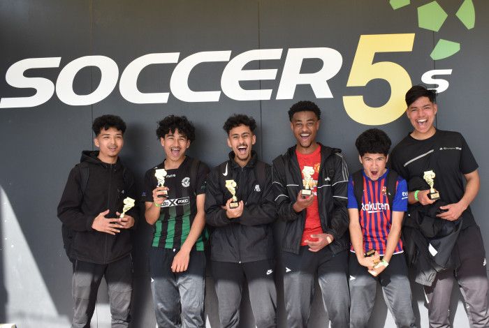 6 teenage boys standing in front of wall smiling with Soccer trophies