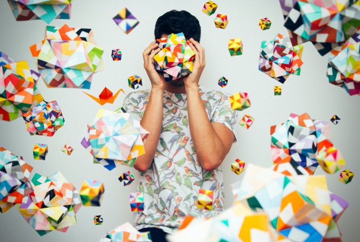 Young person holding origami over their face so you can only see the outline of their face. Rainbow coloured origami all over the wall behind them.