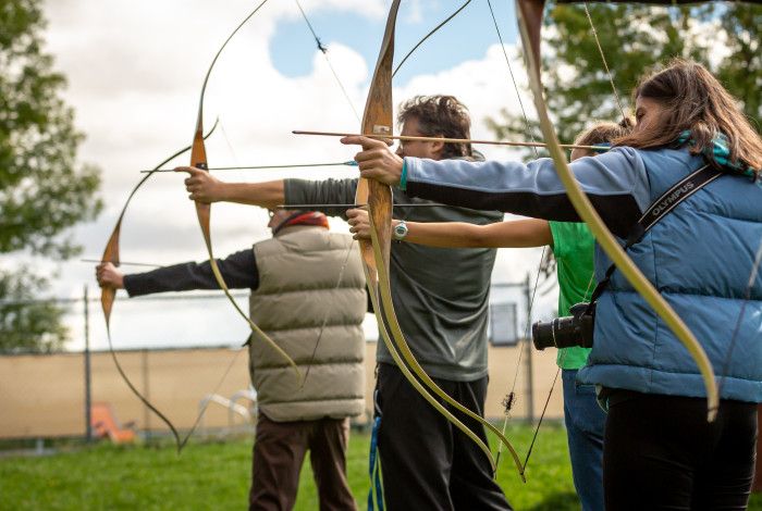 three young people lined up holding bow and arrows aiming to hit a target