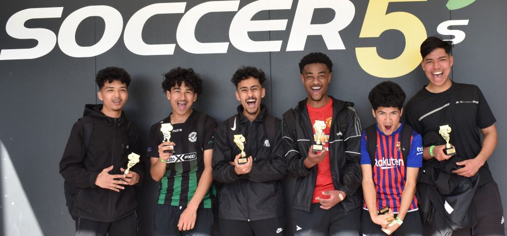 6 teenage boys standing in front of wall smiling with Soccer trophies