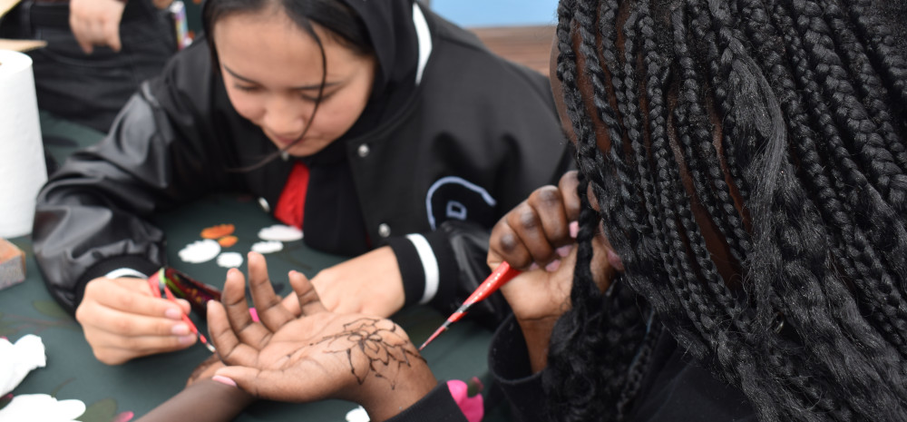 Young person using a henna pen to draw mandala pattern on their hand. Another young person in the background drawing henna art on another young persons arm.