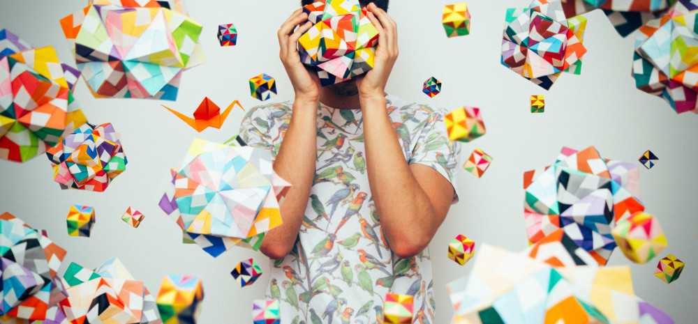 Young person holding origami over their face so you can only see the outline of their face. Rainbow coloured origami all over the wall behind them.